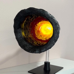 May include: A unique sculpture depicting a cross-section of a celestial body, possibly a planet or star. The outer layer is textured black, encasing vibrant layers of red, yellow, and orange. The sculpture is mounted on a clear acrylic stand with a black base.