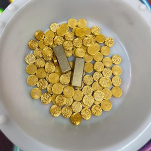 May include: A white bowl filled with gold-colored round discs and two rectangular gold-colored bars.