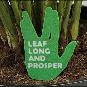 May include: A green plant stake shaped like a hand, with the white text "LEAF LONG AND PROSPER". The stake is in a pot of soil, surrounded by green plants.