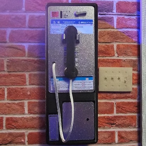 以下が含まれることがあります： 赤いレンガの壁に取り付けられたヴィンテージ公衆電話。 電話は黒とシルバーで、受話器、キーパッド、コインスロットが付いています。 BellSouthのロゴと指示が表示されています。 右側には、ライトスイッチプレートがあります。