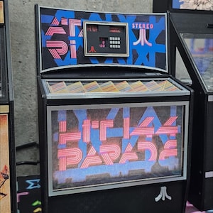 Può includere: Un cabinet da gioco arcade nero e argento con una parte anteriore in vetro. Il cabinet ha una grafica blu e rosa con il testo "STEREO" e "PAC-MAN" nella parte superiore. La parte anteriore in vetro mostra una grafica rosa e blu con il testo "PAC-MAN PARADE".