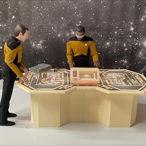 May include: Two figures in yellow and black uniforms stand at a large, octagonal, beige console with a black top. The console has three screens with a starfield background.