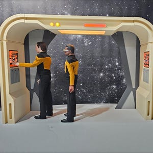 Könnte beinhalten: Zwei Figuren in Starfleet-Uniformen stehen vor einer beige und grauen Tür mit einem beleuchteten Bedienfeld. Die Figuren tragen gelbe und schwarze Uniformen mit einem Starfleet-Abzeichen auf der Brust. Der Hintergrund ist dunkelgrau mit weißen Sternen.
