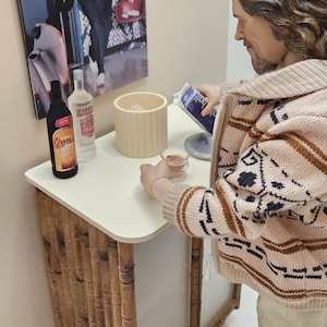 May include: A man wearing a brown and white patterned cardigan stands at a bar with a white countertop and bamboo base. He is pouring a clear liquid from a bottle into a glass. There are two bottles on the counter, one with a clear liquid and one with a dark liquid. There is a white bowl on the counter with a white ice bucket.