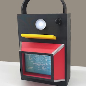 May include: A black, rectangular box-shaped object with a curved handle. It features a yellow shelf, a red compartment with a blueprint image, and a white light. A black knob is on the top.