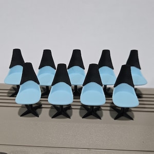 Può includere: Una collezione di sedie in miniatura con sedili azzurri e schienali e gambe neri. Le sedie sono disposte in fila su una superficie grigia, con uno sfondo bianco.