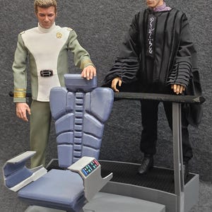 Puede incluir: Dos figuras se encuentran de pie frente a una silla gris y azul con un panel de control. La figura de la izquierda lleva un uniforme verde y blanco. La figura de la derecha lleva una túnica negra con forro morado y texto blanco.