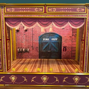 May include: A miniature theater stage with a burgundy and gold frame. The stage features a red brick backdrop, wooden floor, and a burgundy curtain. The words "FIRE EXIT" are visible on a blue door.