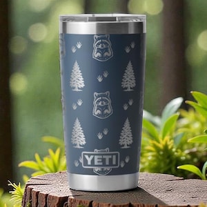 May include: A blue insulated tumbler with a silver rim and base, featuring a pattern of raccoons and pine trees. The tumbler has the brand name "YETI" at the bottom. The tumbler is sitting on a tree stump.