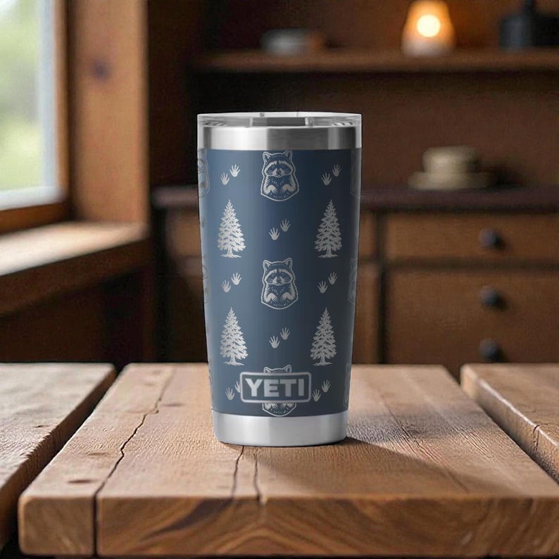 May include: A stainless steel tumbler with a navy blue finish. The tumbler features a pattern of raccoons, pine trees, and the brand name "YETI". The tumbler is designed to keep beverages cold or hot.