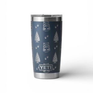 May include: A stainless steel tumbler with a navy blue finish. The tumbler features a repeating pattern of raccoons, pine trees, and paw prints. The brand name "YETI" is displayed at the bottom.