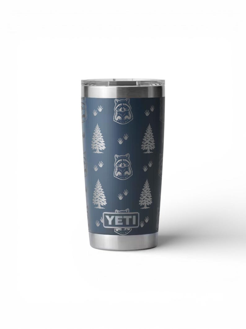 May include: A stainless steel tumbler with a navy blue finish. The tumbler features a repeating pattern of raccoons, pine trees, and paw prints. The brand name "YETI" is displayed at the bottom.