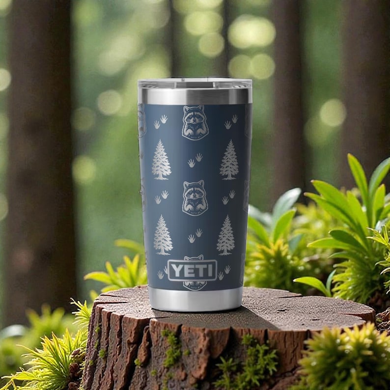 May include: A blue insulated tumbler with a silver rim and base, featuring a pattern of raccoons and pine trees. The tumbler has the brand name "YETI" at the bottom. The tumbler is sitting on a tree stump.