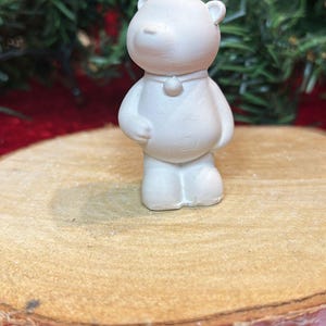 Könnte beinhalten: Kleine, cremeweiße Bärenfigur mit rundem Körper, kleinen Ohren und einem einfachen Kragen. Der Bär steht auf einer runden Holzscheibe, mit einem verschwommenen grünen Girlande im Hintergrund. Ein bezaubernder Dekorationsartikel.