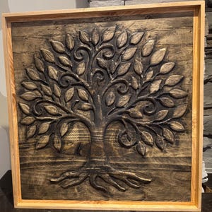 May include: A square wooden wall art piece with a detailed relief carving of a tree. The tree has a dark brown trunk and branches with many leaves. The artwork is framed in light-coloured wood.