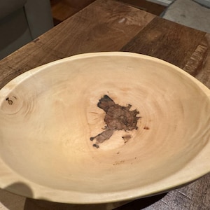 May include: An oval wooden bowl in a light colour, showing the natural wood grain. The bowl has a dark, knotty wood pattern in the centre. It is placed on a wooden surface.