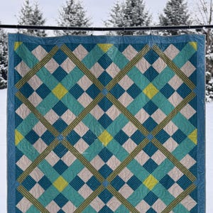 May include: A blue, green, and white quilt with a geometric pattern of squares and stripes. The quilt is hanging on a clothesline in front of a snowy background.