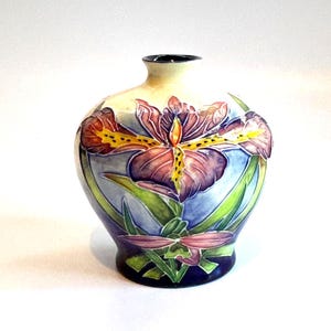 May include: A ceramic vase with a floral iris design. The vase features a cream-colored top, a dark blue base, and a central design of purple, yellow, and orange irises with green leaves. The vase is approximately 6 inches tall.