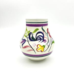 Könnte beinhalten: Eine weiße Keramikvase mit einem Hahn- und Blumendesign. Die Vase hat ein lila Band und grüne und lila Streifen an der Oberseite. Der Hahn ist blau und grün, und die Blumen sind rot, gelb und lila.