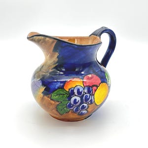 Puede incluir: Jarra de cerámica con asa y borde azules, con un diseño de frutas pintado a mano que incluye uvas, manzanas y limones. La jarra tiene una base marrón y un cuerpo azul, creando una pieza colorida y decorativa.