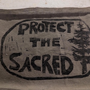 May include: A grey fabric patch featuring the words "PROTECT THE SACRED" in black lettering within a black oval. A black tree illustration is on the right. The patch has a raw, unfinished edge.