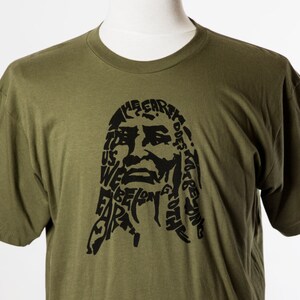 May include: A green t-shirt with a black graphic print of a face made of words. The words include "The Earth Does Not Belong To Us, We Belong To The Earth".