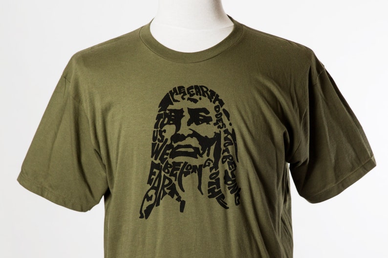 May include: A green t-shirt with a black graphic print of a face made of words. The words include "The Earth Does Not Belong To Us, We Belong To The Earth".
