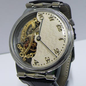 May include: A wristwatch with a silver-toned circular case and a black leather strap. The dial features a cream-colored section with black Arabic numerals and a textured pattern, alongside an exposed gold-toned skeletonized mechanical movement.