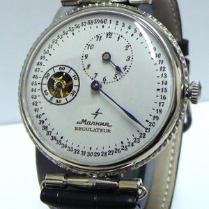 May include: A silver-toned Malnia Regulator watch with a white face and black leather band. The watch features a subdial at the 9 o'clock position and a seconds dial at the 6 o'clock position. The watch has a black leather band with silver-toned hardware.