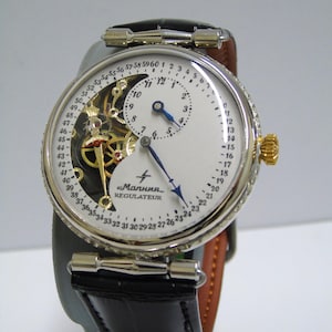 May include: A silver-toned wristwatch with a black leather strap, featuring a white dial with a 60-minute outer track, a sub-dial for hours, and an exposed mechanical movement. The dial displays the text "Малния" and "REGULATEUR" with blue hands.