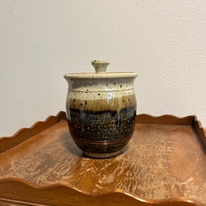May include: A ceramic jar with a lid, featuring a speckled beige top and a dark brown base. The jar has a rounded shape and sits on a wooden surface. The jar is likely used for storage.