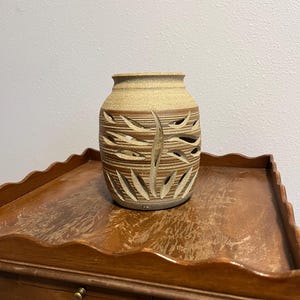 May include: A ceramic vase with a textured, light brown exterior and a darker brown band with leaf-shaped cutouts. The vase sits on a wooden table with a scalloped edge.