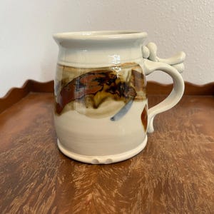 May include: A handmade ceramic mug with a unique handle design. The mug features a cream-colored base with brown and rust-colored abstract patterns. The handle has an artistic, sculptural shape. The mug is sitting on a wooden tray.