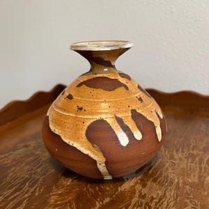May include: A small, brown ceramic vase with a white glaze. The vase has a round body and a narrow neck. The glaze has a drip effect, creating a unique pattern.