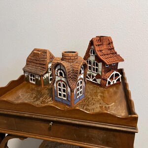 May include: Three ceramic miniature houses with detailed architectural features. The houses have brown, red, and white accents, with textured roofs and window details. They are arranged on a wooden table.