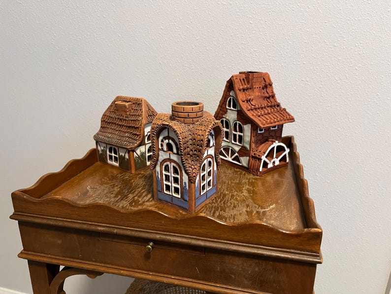 May include: Three ceramic miniature houses with detailed architectural features. The houses have brown, red, and white accents, with textured roofs and window details. They are arranged on a wooden table.