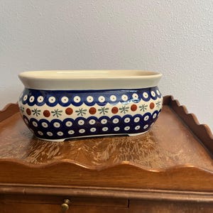 May include: An oval ceramic planter featuring a cream-colored interior and a dark blue exterior decorated with repeating patterns of blue circles, small red dots, and light green star-like shapes arranged in horizontal bands.