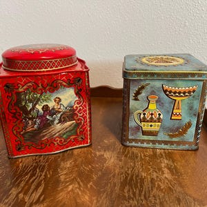 May include: Two vintage decorative metal tins. The red tin features a painting of people in a landscape. The blue tin has a design of a vase and bowl. Both tins have gold accents and lids.
