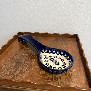 May include: A blue and white ceramic spoon rest with a floral pattern. The spoon rest has a deep blue handle and a bowl-shaped rest with a cream-colored interior. The interior is decorated with blue and dark red floral designs.