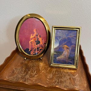 May include: Two framed art prints on a wooden table. One is oval with a gold frame, featuring figures on a red background. The other is rectangular with a gold frame, depicting a woman in a landscape. The table has a decorative, patterned surface.