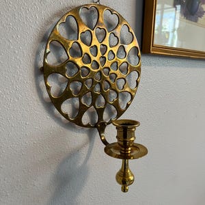 May include: A brass wall-mounted candle holder. The holder features a circular design with heart-shaped cutouts and a single candle cup. The candle holder is attached to a wall with a small black wire.