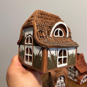 May include: A miniature ceramic house with a brown tile roof, white window frames, and brown trim. The house has multiple windows and a decorative design. The overall color scheme is brown, white, and green.