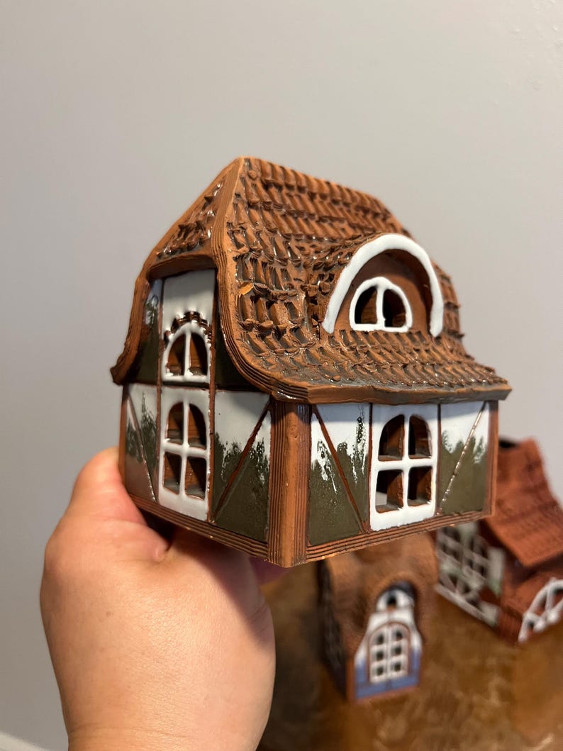 May include: A miniature ceramic house with a brown tile roof, white window frames, and brown trim. The house has multiple windows and a decorative design. The overall color scheme is brown, white, and green.