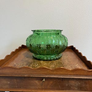 May include: A round, emerald green glass bowl featuring a textured, ribbed exterior pattern with a scalloped base and a wide, open top. The glass has a translucent quality, catching the light to highlight the vertical ridges and pebbled surface details across the body of the vessel.
