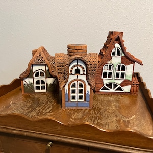 May include: Three ceramic house figurines with detailed brown roofs, white window frames, and colorful accents. The houses have unique architectural designs, including chimneys and varying window shapes. They are displayed on a wooden surface.