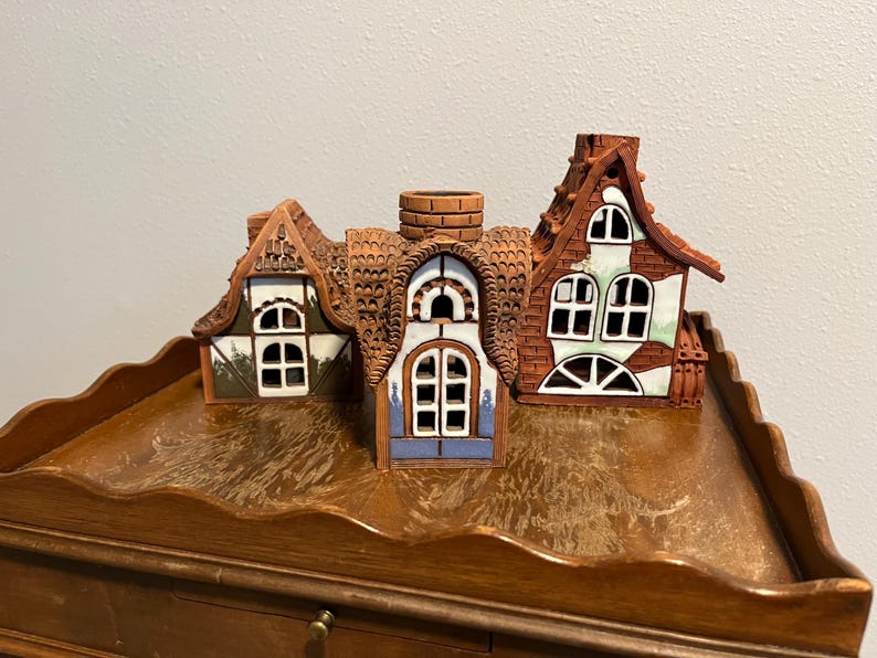 May include: Three ceramic house figurines with detailed brown roofs, white window frames, and colorful accents. The houses have unique architectural designs, including chimneys and varying window shapes. They are displayed on a wooden surface.