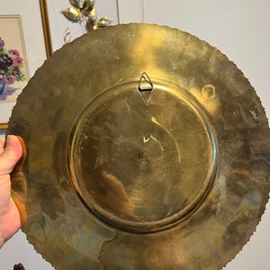 May include: A large, round brass plate with a decorative, scalloped edge. The back of the plate features a central recessed area and a triangular hanger. The plate has a warm, golden hue and shows signs of age.