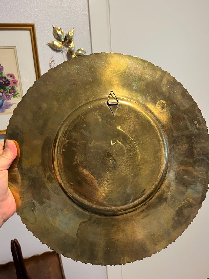 May include: A large, round brass plate with a decorative, scalloped edge. The back of the plate features a central recessed area and a triangular hanger. The plate has a warm, golden hue and shows signs of age.