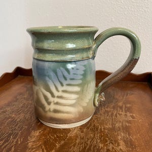 May include: Handmade ceramic mug with a green rim and handle, transitioning to blue and tan tones on the body. The mug features a fern leaf design and is suitable for hot or cold beverages.