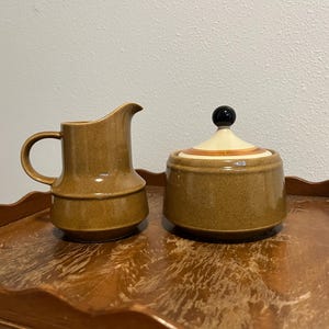 May include: A ceramic creamer and a lidded sugar bowl with a speckled brown glaze. The creamer features a curved handle and a pouring spout. The sugar bowl has a matching brown base, a cream-colored lid with orange circular bands, and a black knob.